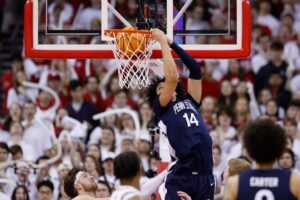 The cover for this college basketball picks today article shows Yanic Konan Niederhauser #14 of the Penn State Nittany Lions scoring on a reverse dunk during the second half of the game against the Wisconsin Badgers.
