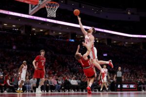 The cover for this college basketball picks today article shows Brandon Noel #14 of the Ohio State Buckeyes colliding with Sam Hoiberg #1 of the Nebraska Cornhuskers while shooting the ball.