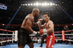 A wide-angle action shot of Floyd Mayweather Jr. in black trunks and Manny Pacquiao in red trunks boxing in a professional ring. Mayweather is throwing a punch while Pacquiao defends. The full ring, ropes, referee, and arena lights are visible.